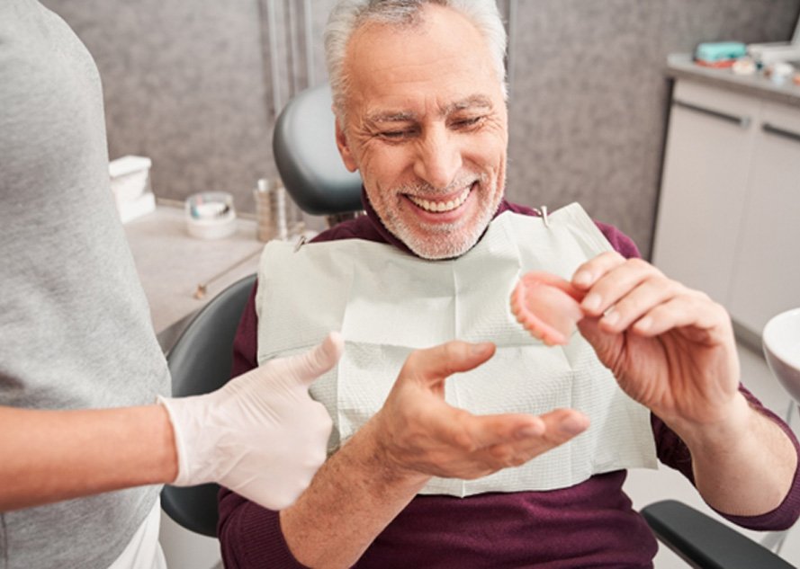 Man holding a denture in the dentist’s office and smiling