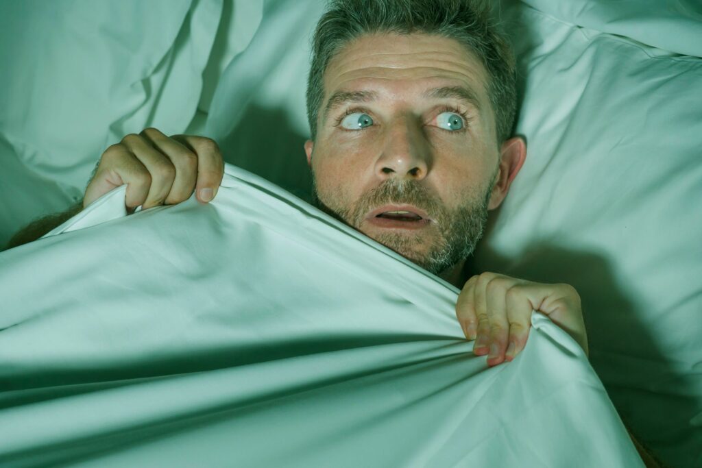 Man pulling bedsheets to face looking away in fear